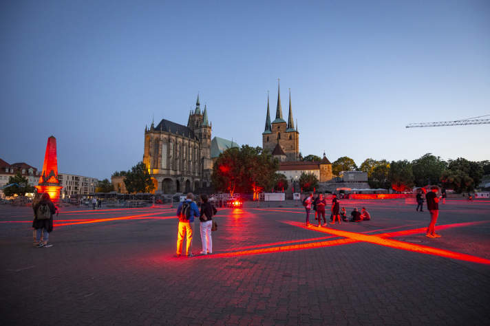 Rotes Licht wird auf einen großen Platz in Streifenform projiziert, im Hintergrund zwei Kirchengebäude.