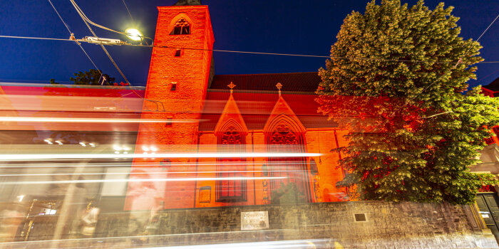 Eine Kirche wird rot angestrahlt, davor sieht man Schlieren von Licht einer vorbeifahrenden Straßenbahn.