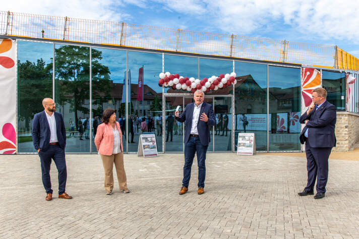 Vier Personen stehen vor einem mit Luftballon und Bannern geschmückten Glasbau.