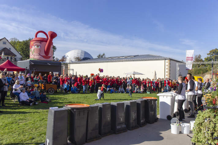 eine Reihe von Mülltonnen, an denen eine Gruppe Menschen "Musik" macht, davor Publikum auf einer Wiese