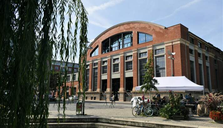 Backsteingebäude vor einem Platz mit Baum, auf dem ein kleiner Pavillon aufgebaut ist.