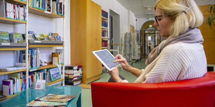 Frau sitz in Bibliothek vor Tisch mit Zeitschriften und schaut auf ihr Tablet 