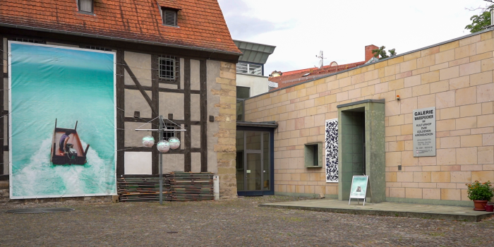 Außenansicht zweier Gebäude, an einem hängt ein großes blaues Banner.