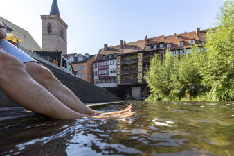 Vor der Krämerbrücke hält ein Mann an einem heißen, sonnigen Tag seine Beine in einen Fluss.
