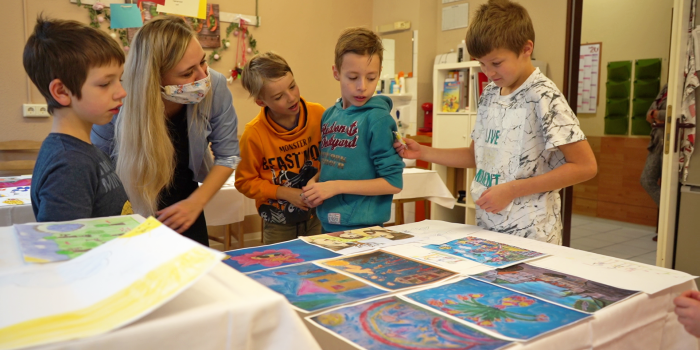 Foto: Titelauswahl Familienpass 2021 © Stadtverwaltung Erfurt Vier Kinder und eine Erwachsene stehen um gemalte Bilder herum die auf einem Tisch liegen.
