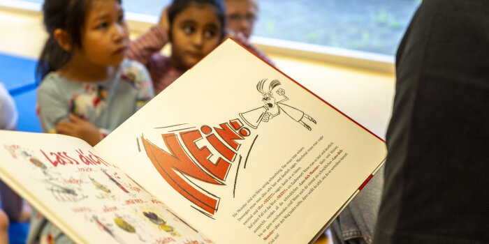 In einem aufgeschlagenen Buch steht groß „Nein“. Im Hintergrund sind drei Kinder zu erkennen. 