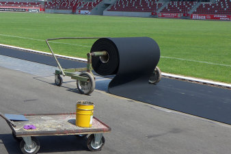 Foto: Tartanbahn Kunststoffbelag © Erfurter Sportbetrieb In einem Stadion wird auf einem Asphaltboden ein schwarzer Kunststoffbelag ausgerollt.