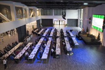 Ein großer, moderner Saal mit vielen Personen, die an Tischen sitzen und einem Redner zuhören.