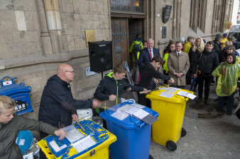 Eine Trommelgruppe spielt auf gelben und blauen Mülltonnen, der OB und der MP hören zu 