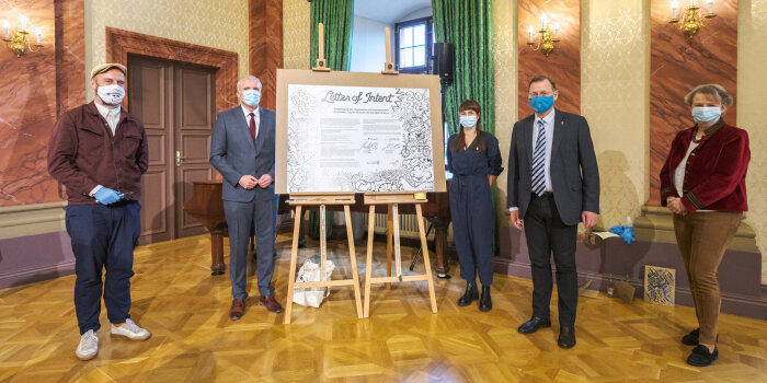Ministerpräsident, Oberbürgermeister und drei Personen vor einer großen Urkunde im Festsaal der Staatskanzlei. 