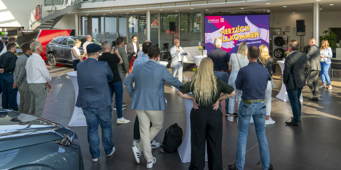 ein Mann spricht vor Publikum im Foyer eines Autohauses, im Hintergrund Arbeitsplätze und Fahrzeuge