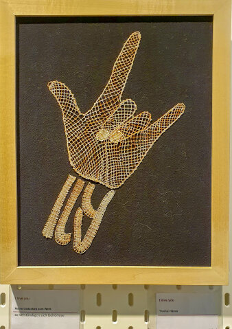 in einem Rahmen ist eine aus goldfarbenen Fäden geklöppelte Hand abgebildet, die Mitte- und Ringfinger einknickt