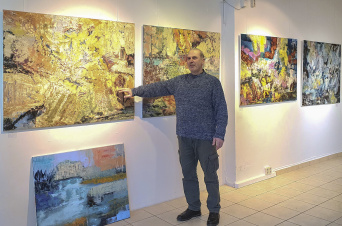 ein Mann im blauen Pullover zeigt auf eins von mehreren bunten Gemälden, die an einer Wand hängen