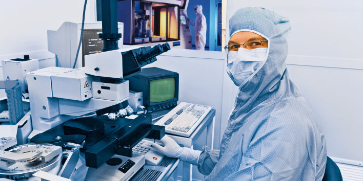 Ein Mann im Reinraumanzug und mit Mundschutz blickt vom Mikroskop auf in die Kamera