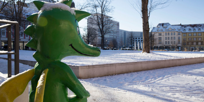 Foto: Hirschgarten Tabaluga Winter © Stadtverwaltung Erfurt Ein Blick über die Schulter der Tabaluga-Figur auf den zugeschneiten Hirschgarten, im Hintergrund zu sehen sind Plattenbauten und alte Gebäude.
