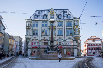 Zentraler Blick auf den alten Angerbrunnen, zu dessen Seite die Gleise der Straßenbahn verlaufen, auf den Dächern und dem Gehweg liegt eine leichte Schneedecke.