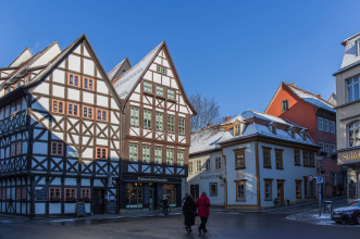 Zu sehen sind alte Fachwerkhäuser mit Schnee auf den Dächern, davor ein Platz, auf dem sich wenige Menschen befinden und der leicht schräge Aufgang zur engen Gasse der Krämerbrücke.