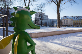 Hirschgarten Tabaluga Winter Ein Blick über die Schulter der Tabaluga-Figur auf den zugeschneiten Hirschgarten, im Hintergrund zu sehen sind Plattenbauten und alte Gebäude.