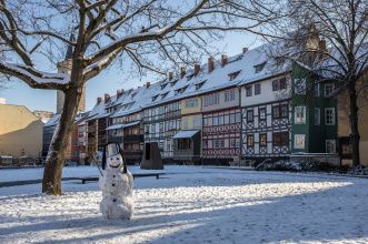 Krämerbrücke Schneemann Im Vordergrund ist ein Schneemann mit einem Metalleimer auf dem Kopf zu sehen, stehend auf einer verschneiten Wiese mit kahlen Bäumen. Im Hintergrund die Häuser der Krämerbrücke von außen mit ihren zugeschneiten Dächern.