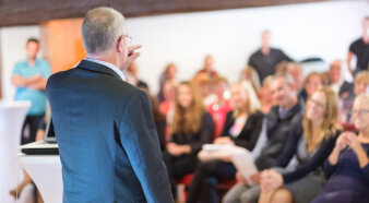 Foto: Redner vor Publikum © kasto/Fotolia Älterer Mann hält vor einem verschwommen dargestellten Publikum einen Vortrag.