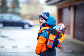 Ein kleiner Junge mit Schulrucksack und Winterlklamotten steht im Winter vor einer Garage und schaut über die linke Schulter in die Kamera.