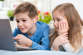 Zwei Kinder schauen auf einen Laptop und haben je ein Teil eines In-Ear Kopfhörerkabels im Ohr.