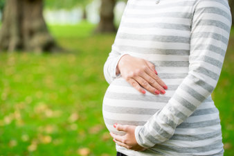 Eine schwangere Frau, deren Gesicht nicht zu erkennen ist, hält ihren Bauch sorgsam mit beiden Händen während sie in einem Garten mit großen Bäumen steht.