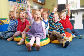 Eine Gruppe von sechs Kindergartenkidnern sitzt auf dem Boden und alle heben mit offenen Mündern enthusiastisch die Hände.