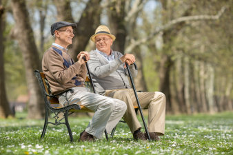 Zwei Senioren sitzen im Park auf einer Bank. Sie haben beide Hüte auf, alterstypische Kleidung an und schwarze Gehstöcke in den Händen.