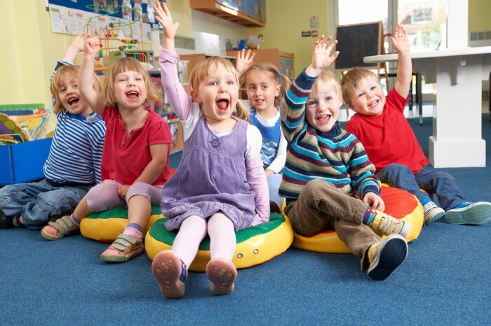 Kindergartengruppe Eine Gruppe von sechs Kindergartenkindern sitzt auf dem Boden und alle heben mit offenen Mündern enthusiastisch die Hände.