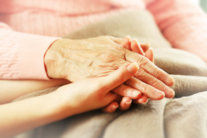 Seniorenpflege Die Hand einer gealterten Frau liegt in der Hand einer jüngeren Frau auf einem Bett.