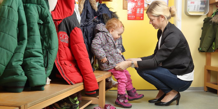 Eine junge Frau im Businessoutfit holt ein kleines Mädchen aus der Kindertagesstätte ab und zieht ihr die Jacke an.