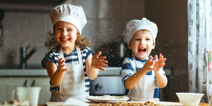 Zwei Kinder mit Schürzem und Kochmützen haben Spaß beim Backen mit Mehl. Sie klatschen und verteilen es überall