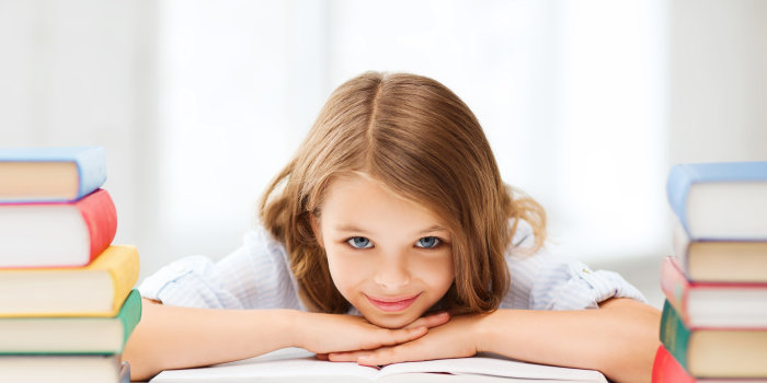 Ein Mädchen im Grundschulalter liegt auf ihre Arme gestützt auf einem Buch und schaut lächelnd in die Kamera. Zu ihren beiden Seiten sind Stapel von Büchern.