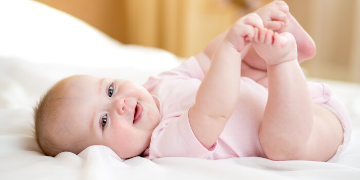 Ein Baby liegt lachend auf einem hellen, weichen Untergrund und fässt sich an die Füße.