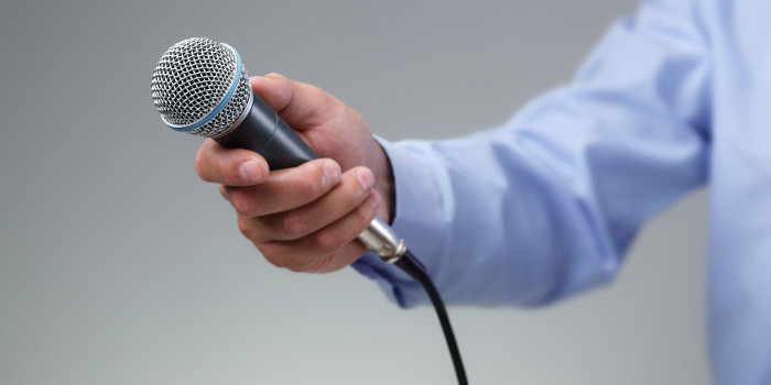 Ein Mikrofon wird von einem Mann, dessen blau gekleideter Arm nur zu sehen ist, Richtung Kamera gehalten.