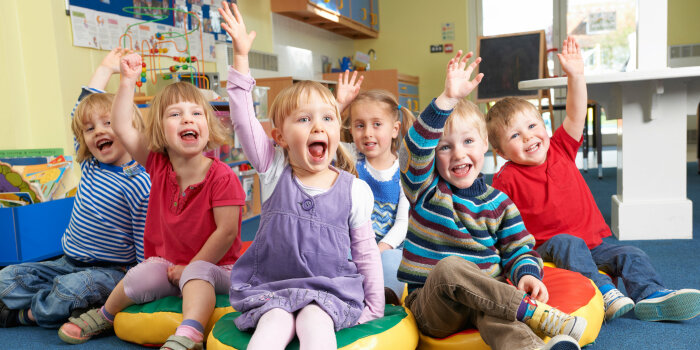 Eine Gruppe von sechs Kindergartenkindern sitzt auf dem Boden und alle heben mit offenen Mündern enthusiastisch die Hände.