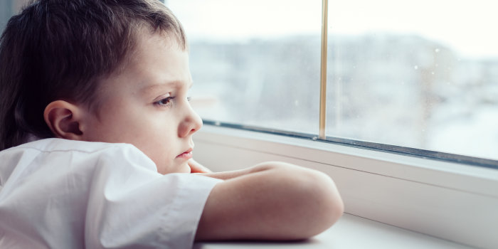 Ein kleiner Junge lehnt auf einem Fensterbrett und schaut mit nachdenklichen und traurigen Gesichtsausdruck aus einem Fenster.
