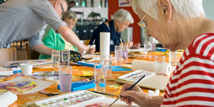 Foto: Malkurs für Senioren © belahoche/Fotolia Mehrere Senioren sitzen an einem Tisch mit Malkästen und bereits vollendeten Bildern vor sich. Ein Helfer geht herum und berät.