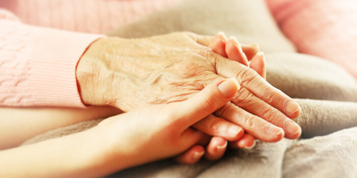 Foto: Seniorenpflege © Africa Studio/Fotolia Die Hand einer gealterten Frau liegt in der Hand einer jüngeren Frau auf einem Bett.