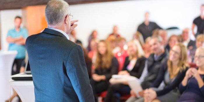 Älterer Mann hält vor einem verschwommen dargestellten Publikum einen Vortrag.