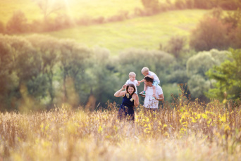 Glückliche Eltern tragen ihre zwei kleinen Kindern auf den Schulter. Sie laufen im Spätsommer durch eine grüne Landschaft.
