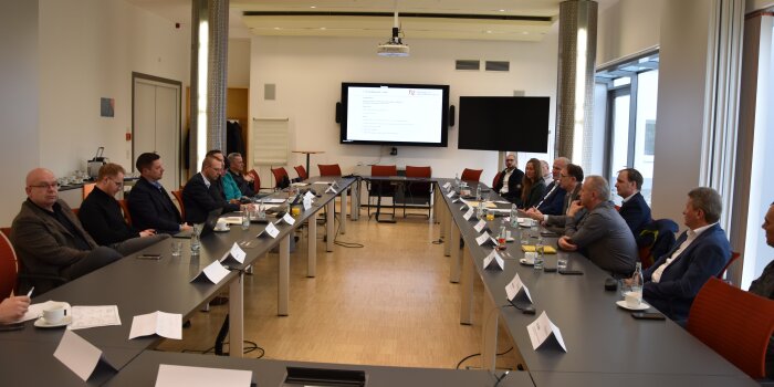 Mehrere Personen sitzen sich an einem Konferenztisch gegenüber. An der Stirnseite hängt ein Monitor an der Wand.  