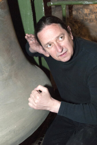Mann mittleren Alters im schwarzen Outfit horcht an einer großem Glocke