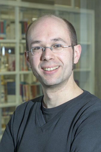 Junger Mann mit Brille, wenig Haar und dunklem Oberteil vor einem Bücherregal stehend