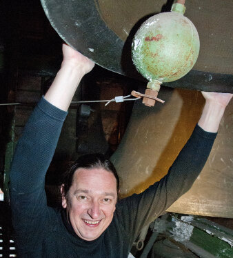 Mann mittleren Alters im schwarzen Outfit hält sich an einer großem Glocke fest