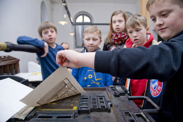Benary-Speicher: Kinder beim Drucken einer eigenen Buchseite Kinder stehen an einer Druckerpresse und drucken eine Buchseite