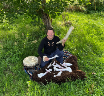 Foto: Arnd Adje Both und seine steinzeitlichen Instrumente © Arnd Adje Both Ein Mann sitzt mit steinzeitlichen Instrumenten auf einer Wiese im Grünen und hält ein Horn in die Höhe