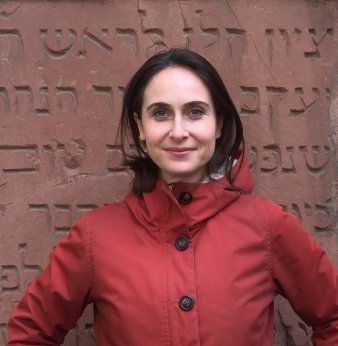 Foto: Naomi Lubrich © N. Lubrich Eine Frau mit dunklen Haaren in rotem Anorak steht vor einer rötlich-braunen Steintafel, auf der hebräische Schriftzeichen eingraviert sind, und blickt in die Kamera.