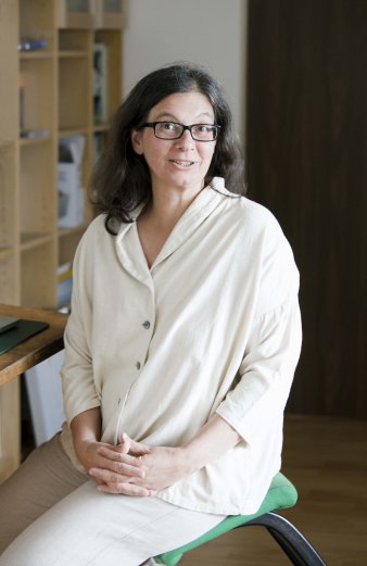 Foto: Marion Steinicke © Henriette Kriese Eine hell gekleidete Frau mit offenen, langen schwarzen Haaren und schwarz umrandeter Brille schaut lächelnd in die Kamera. Sie sitzt auf einem grün bezogenen Hocker. Im Hintergrund sind helle Holzregale sowie am linken Bildrand die Ecke einer Computertastatur auf einem Schreibtisch zu sehen.